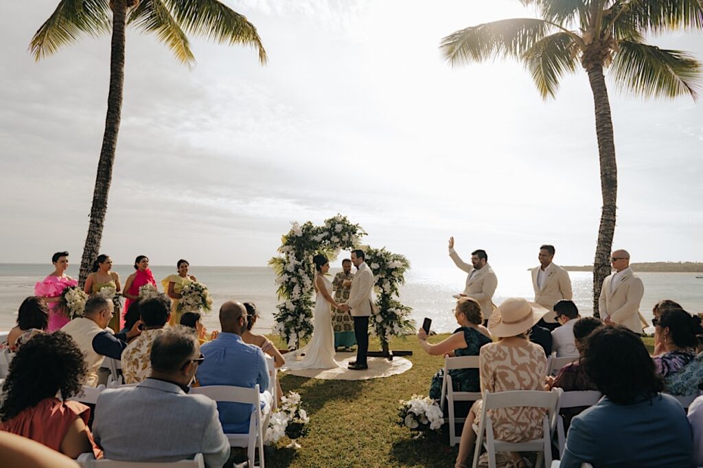 094_bride-groom-at-fiji-wedding-ceremony-intercontinental Wide shot of bride and groom at their Fiji wedding ceremony at Intercontinental, with the groomsman waving his hand after being introduced