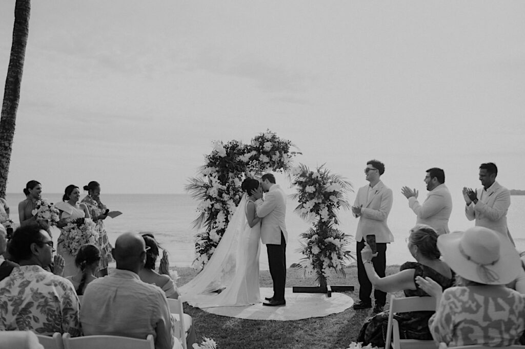 111_couple-share-first-kiss-intercontinental-wedding Wide shot of the couple sharing their first kiss at their Intercontinental wedding in Fiji, with guests clapping in the background. Black and white photo