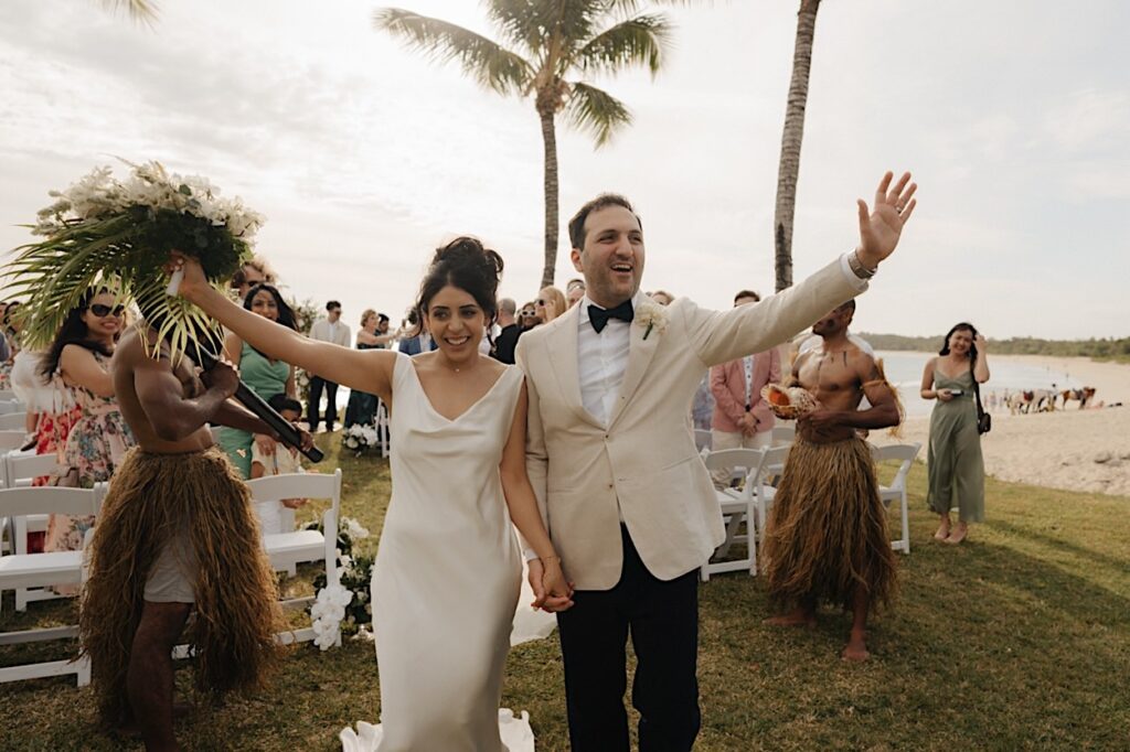 117_couple-celebrate-nuptials-fiji-resort-wedding-coral-coast Bride and groom celebrating their nuptials at a Fiji resort wedding on Coral Coast, cheering with hands in the air as they reach the end of the aisle