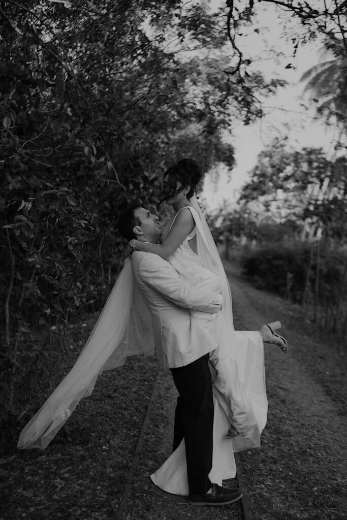 161_bride-groom-portraits-fiji-wedding Black and white portrait of the groom lifting the bride during their Fiji wedding, set against a scenic tropical backdrop