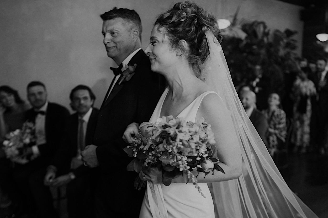 077_bride-entrance-glasshaus-brucke-wedding-melbourne Bride walking down the aisle with her dad on her arm during the Glasshaus Brucke wedding in Melbourne, captured in black and white