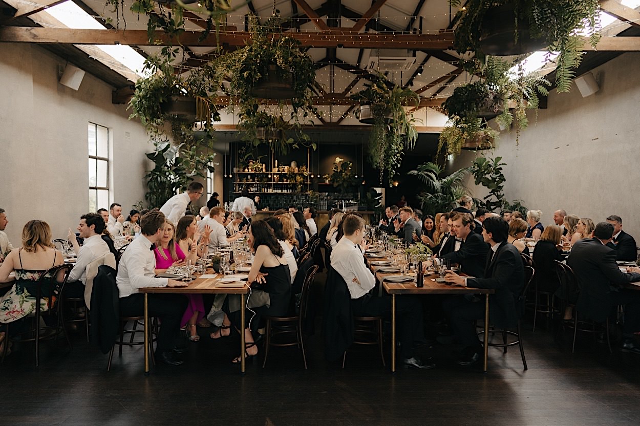 146_guests-reception-glasshaus-brucke-wedding-melbourne Wide shot of guests seated at the reception at Glasshaus Brucke in Melbourne, with lush greenery hanging above