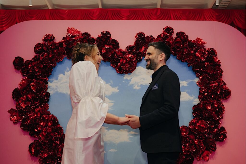 073_couple-laughing-altar-electric-wedding-collingwood Bride and groom laughing while holding hands in front of a colorful love heart backdrop at Altar Electric Wedding in Collingwood