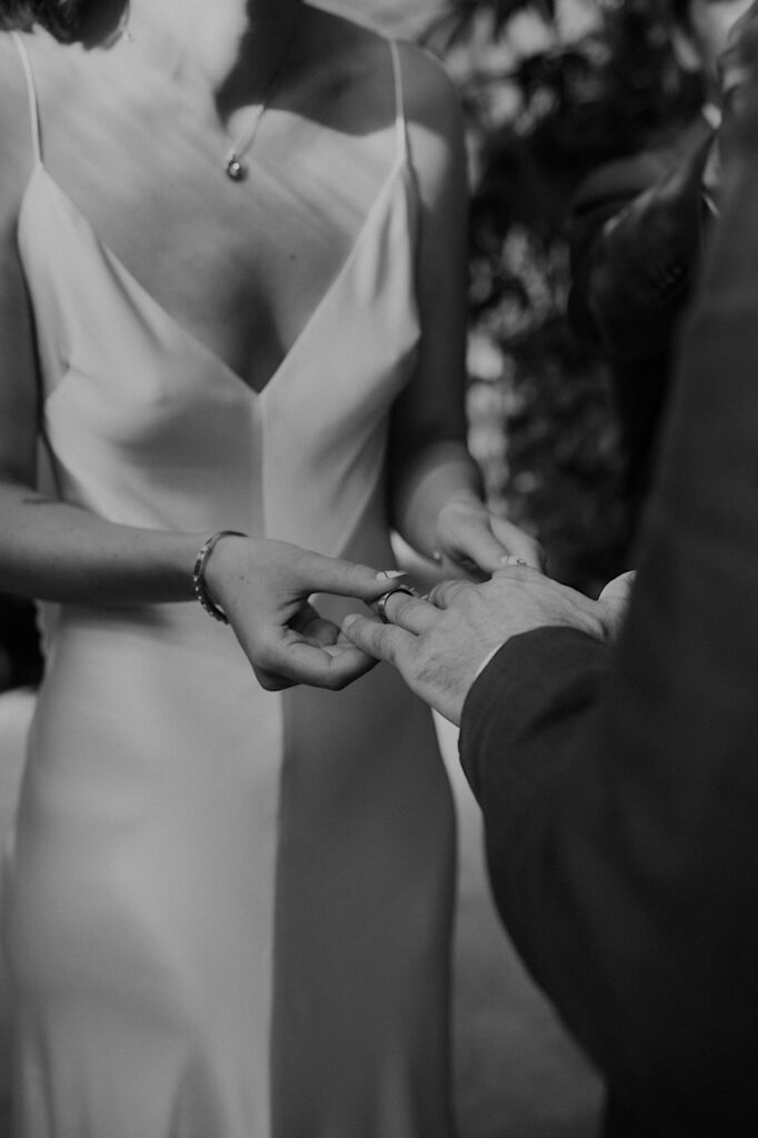 Black-and-white close-up of the bride placing the groom’s ring on his finger during their Glasshaus Inside wedding ceremony